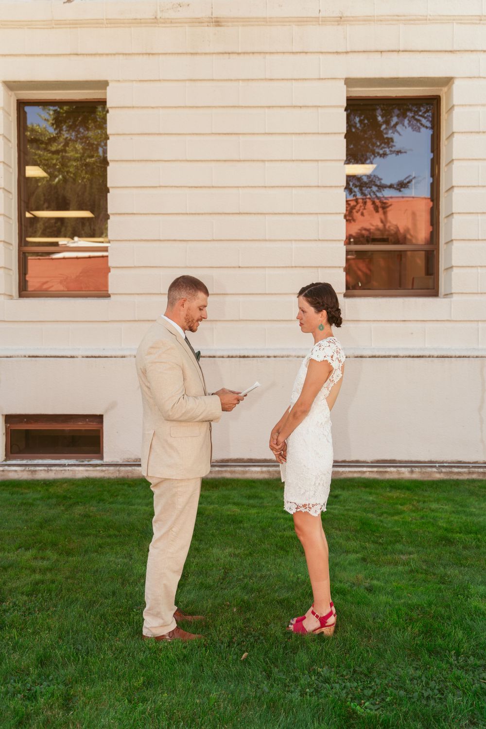 A couple stands on green grass near a white building, exchanging vows in beige and white attire during an outdoor ceremony.