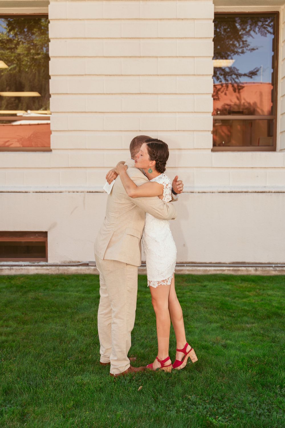 A couple embraces on a lawn in front of a white building, wearing beige and white wedding attire.