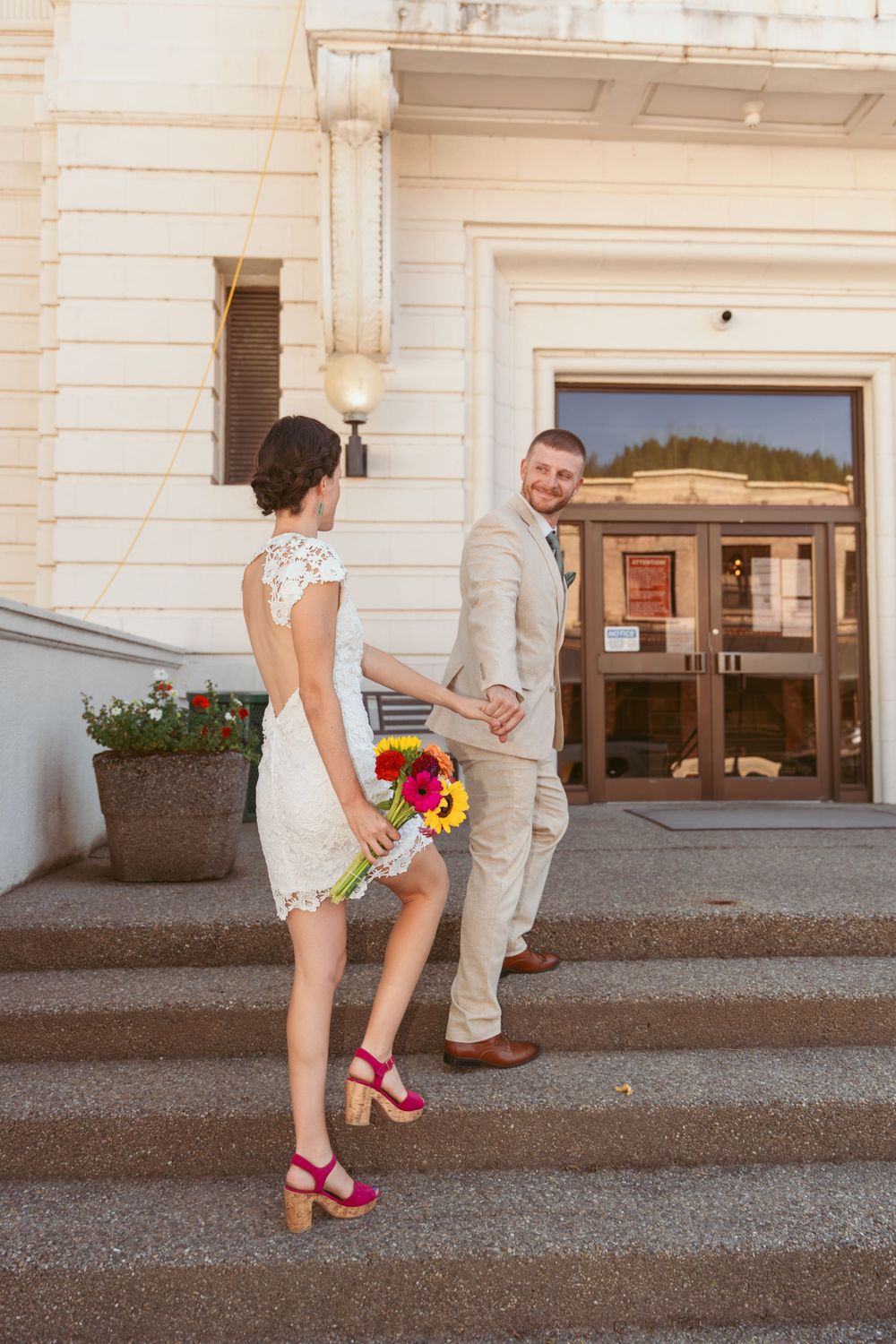 A couple poses playfully on stone steps outside a white building entrance with bright floral bouquet.