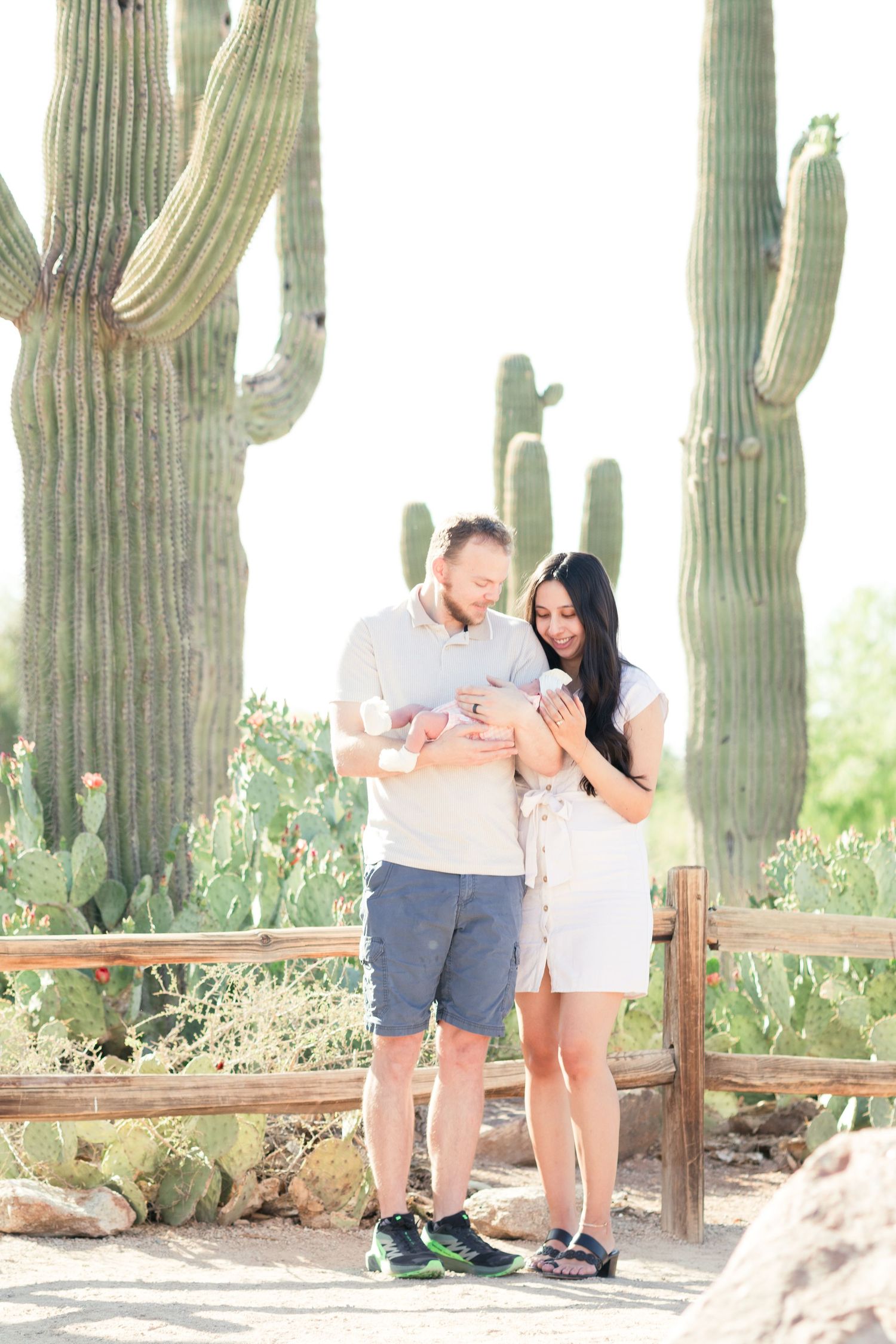 Parents smiling down at newborn daughter in dad's arms in front of a wood fence and cactus at the Gilbert Riparian Preserve.