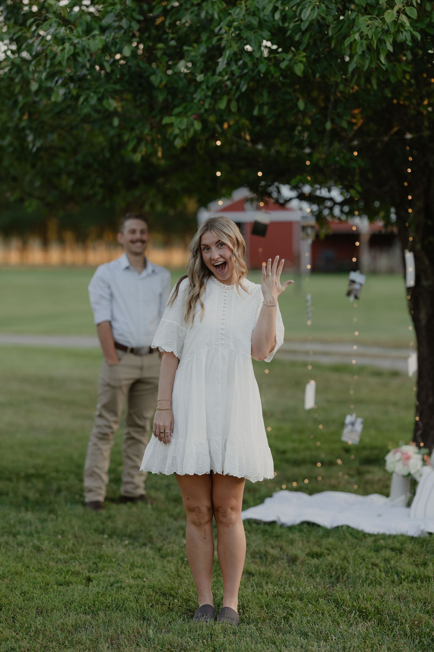 Excited blonde woman in white dress shows off engagement ring while man smiles behind her outdoors.