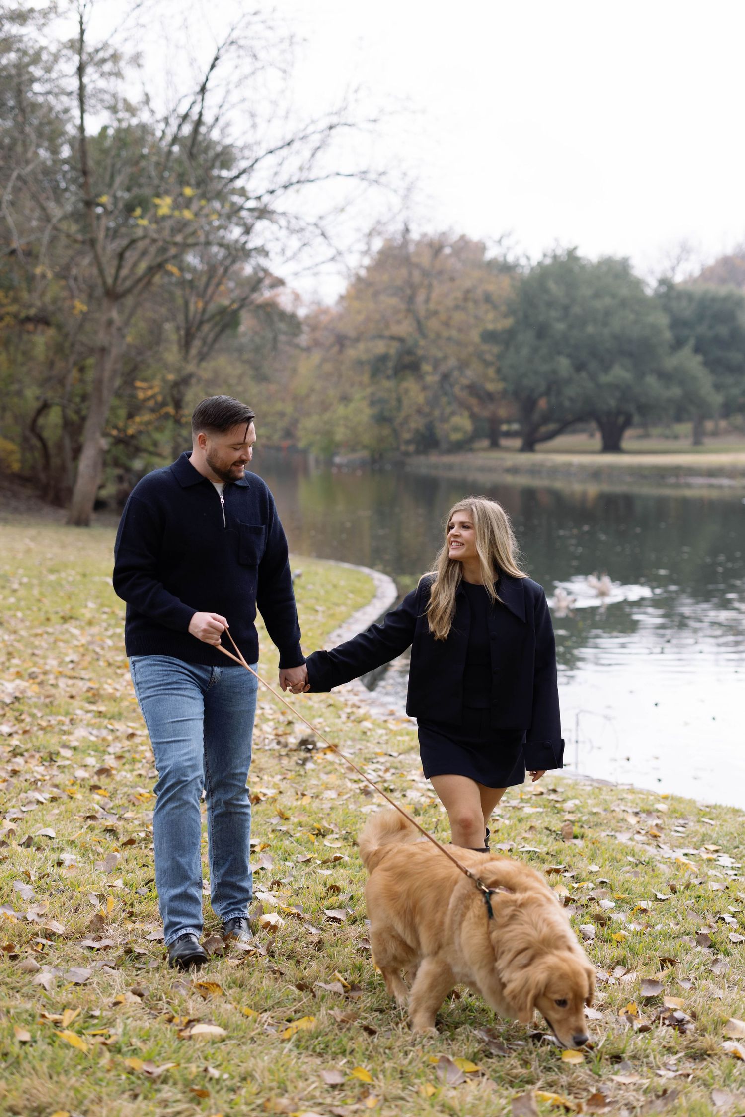 Couple walking their golden retriever on an autumn day by a serene pond with fallen leaves on the ground.