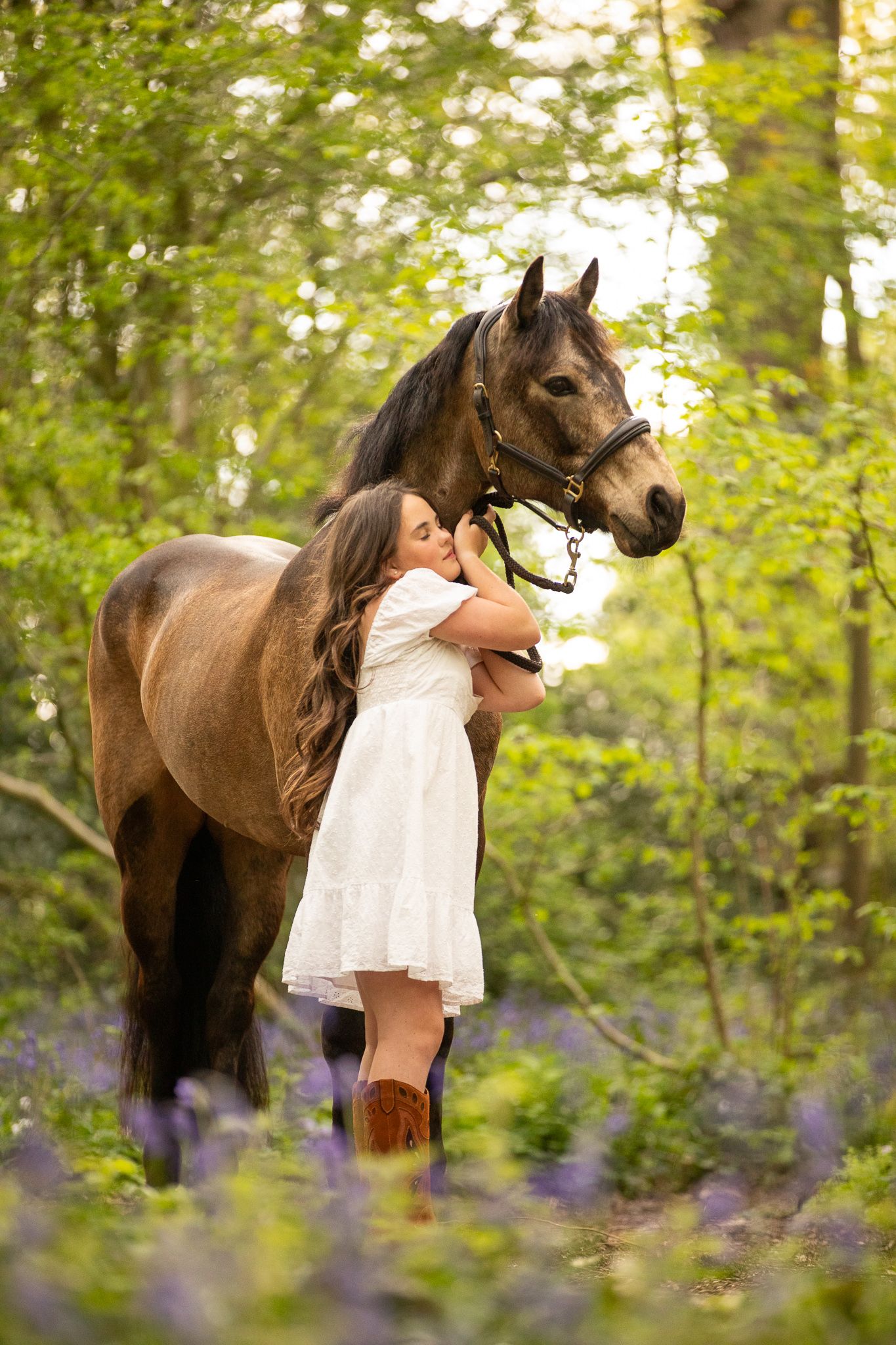 A brown horse and someone in a white dress stand among blooming purple lupines in a sunlit woodland setting.