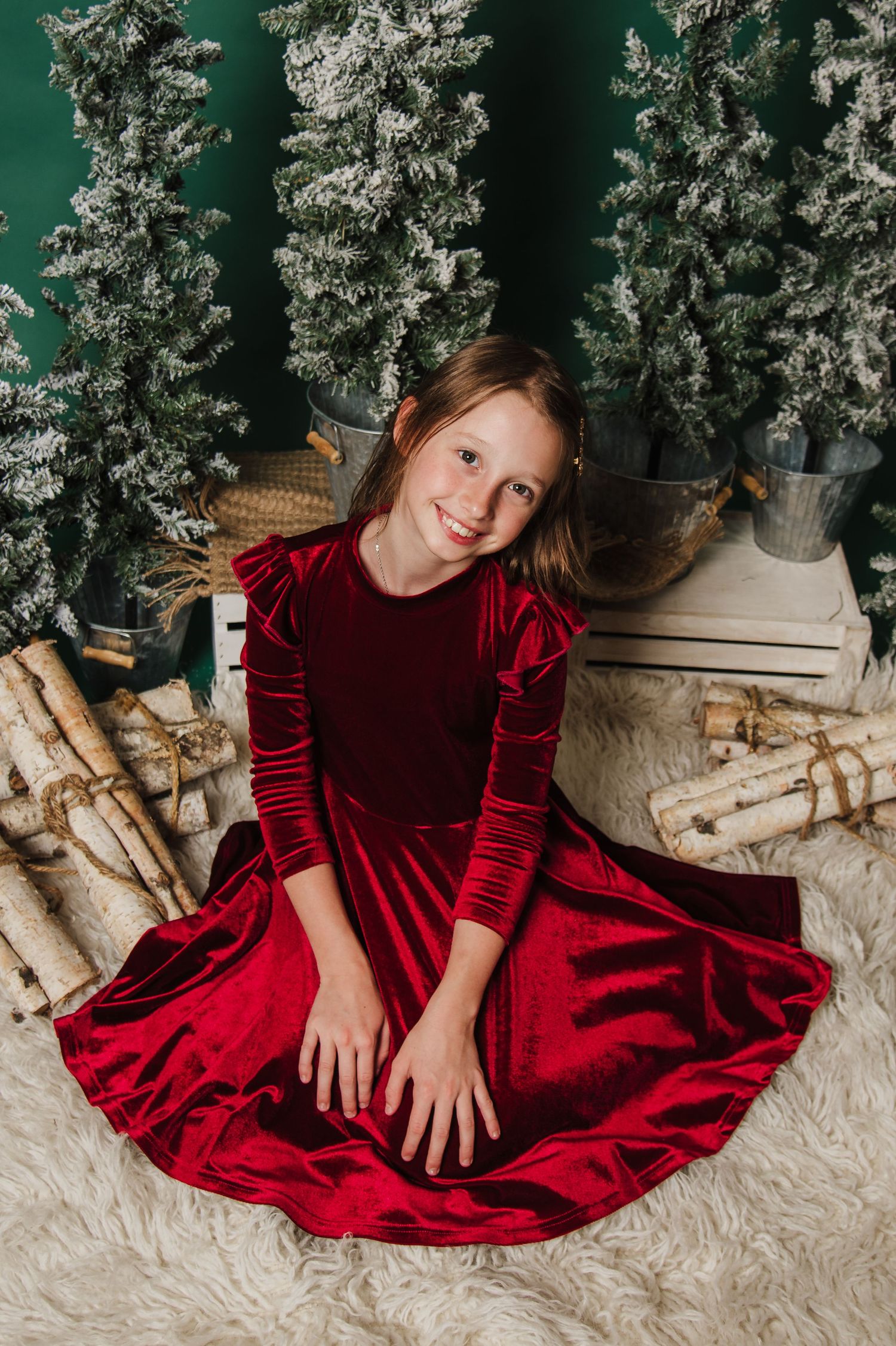 A festive holiday photo session featuring a velvet red dress against a cozy winter backdrop with birch logs and greenery.