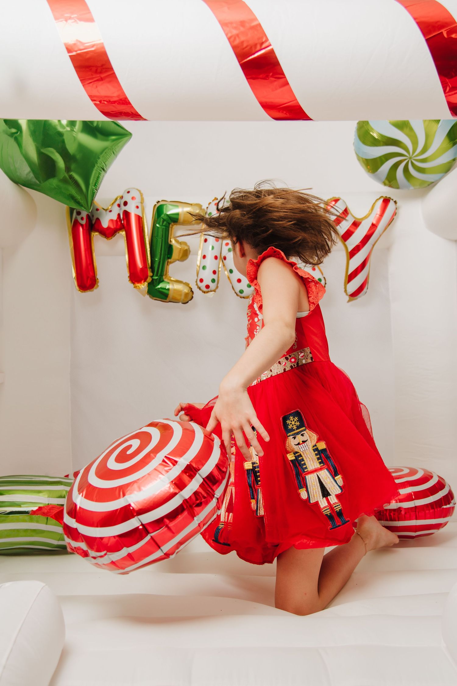 Playful holiday photo shoot with red dress and candy cane balloon decorations on white background.