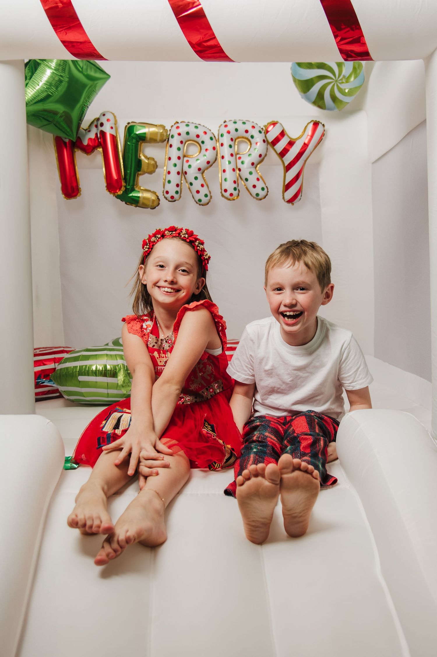 Two children sit barefoot under festive Christmas decorations and a Merry sign.