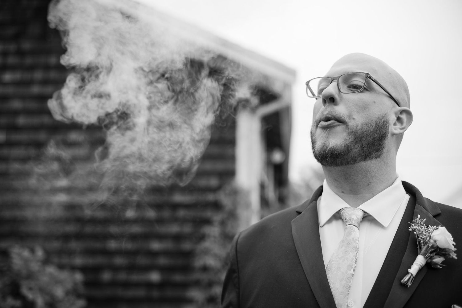 Black and white photo of a person in a suit wearing glasses exhaling smoke against a brick wall backdrop.
