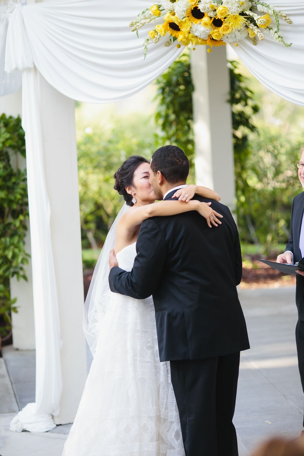 Intimate embrace during wedding ceremony beneath white draped pavilion with yellow floral accents.