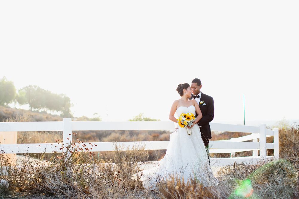 A newly married couple embraces outdoors along a white fence path during golden hour at Summit House in Fullerton.