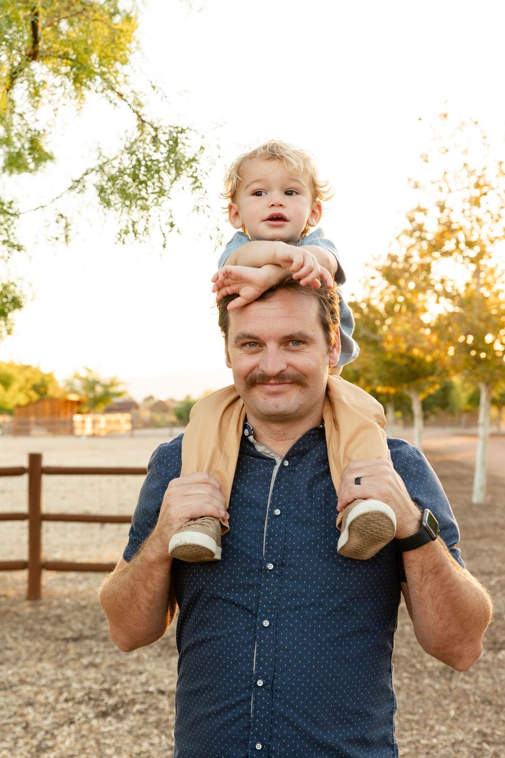 2 year old boy sitting on top of dads shoulders. dad smiling at the camera, boy arms stretched out on top of dads head looking longingly off