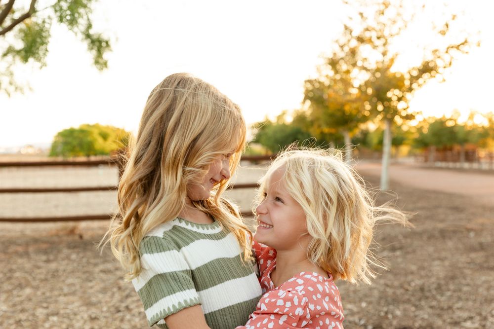 two girls giggling at eachother. 5 and 8 years old. one wearing cream and sage green striped dress. the other a pink polka dot dress