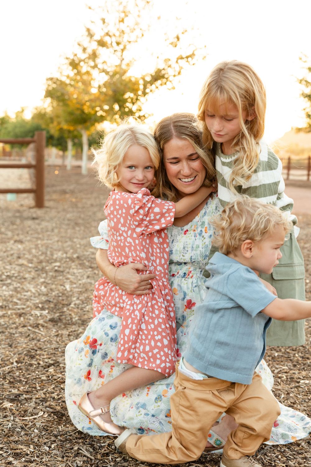 mom in a pretty floral dress kneels down hugging 2 young girls while a 2 year old boy tries to get free of her grasp