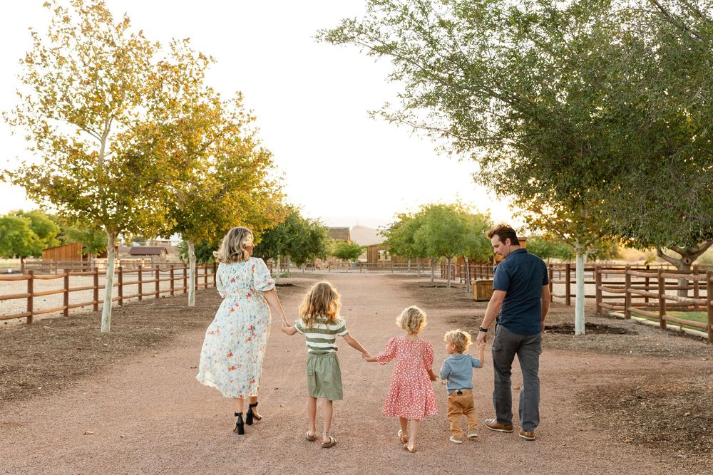family of 5 holding hands walking together away from the camera down the lane with fences on either sides and trees hanging over top