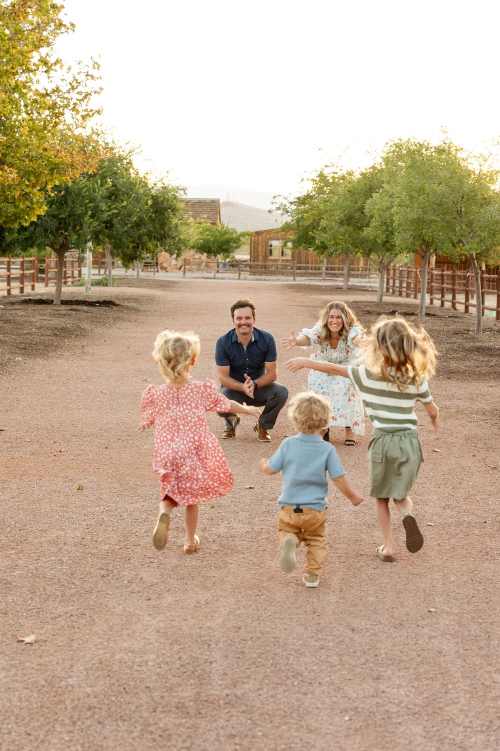a farm land in the background. mom and dad squatting down arms wide open getting ready to receive 3 young  kids running into their arms.