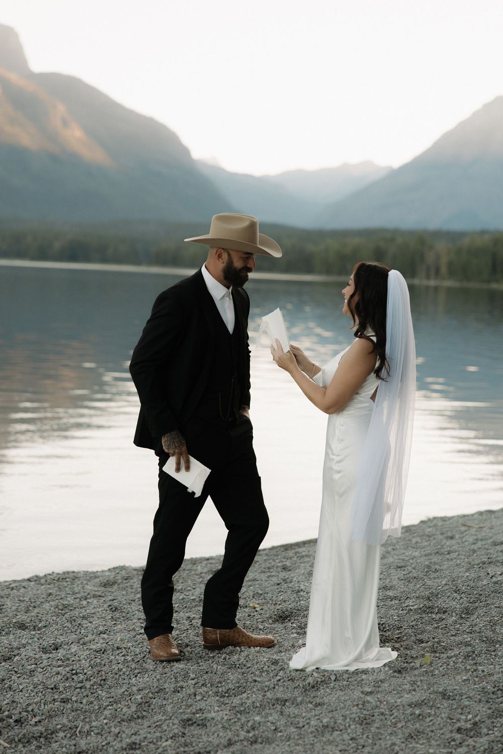 A romantic wedding photoshoot on a lakeside beach with mountains in the background at dusk.