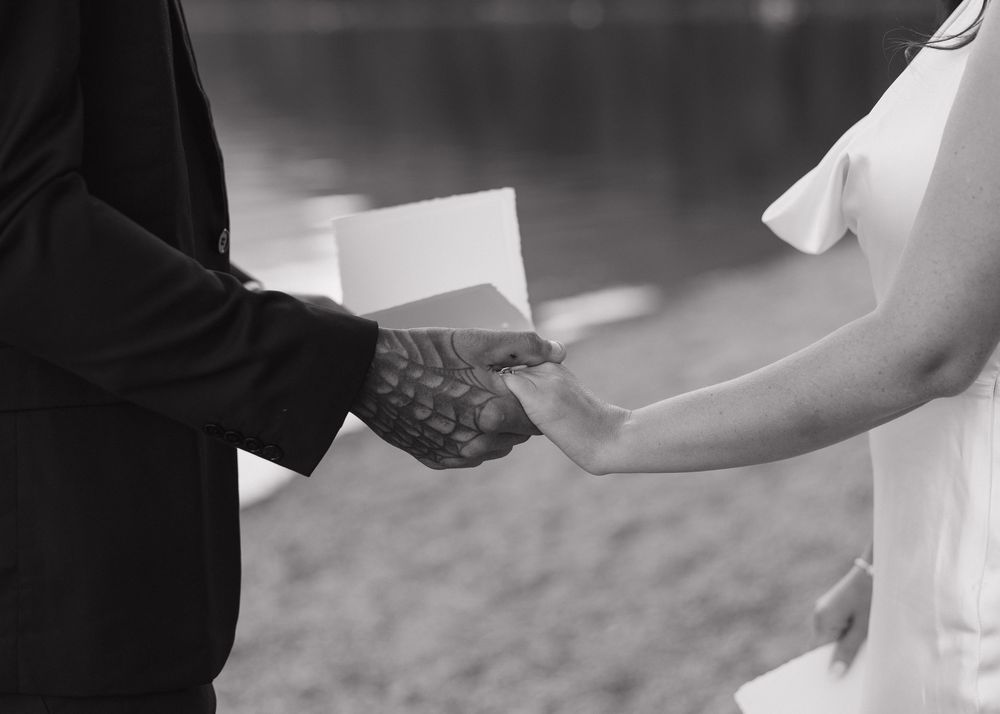 A black and white photograph shows hands exchanging wedding rings during an outdoor wedding ceremony.