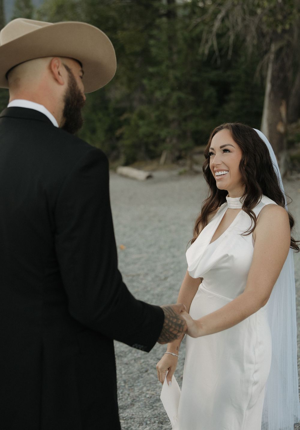 Couple shares a joyful moment during their outdoor wedding ceremony with lush forest backdrop on a rocky beach.