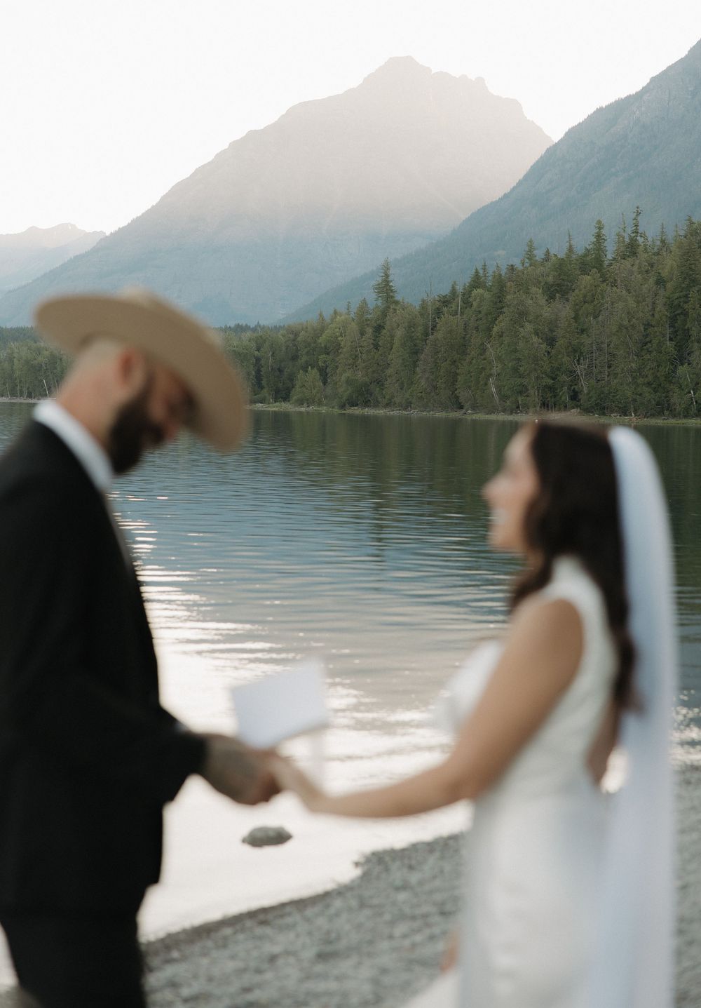 A couple exchanging wedding vows beside a serene lake with mountain views in the background during sunset.