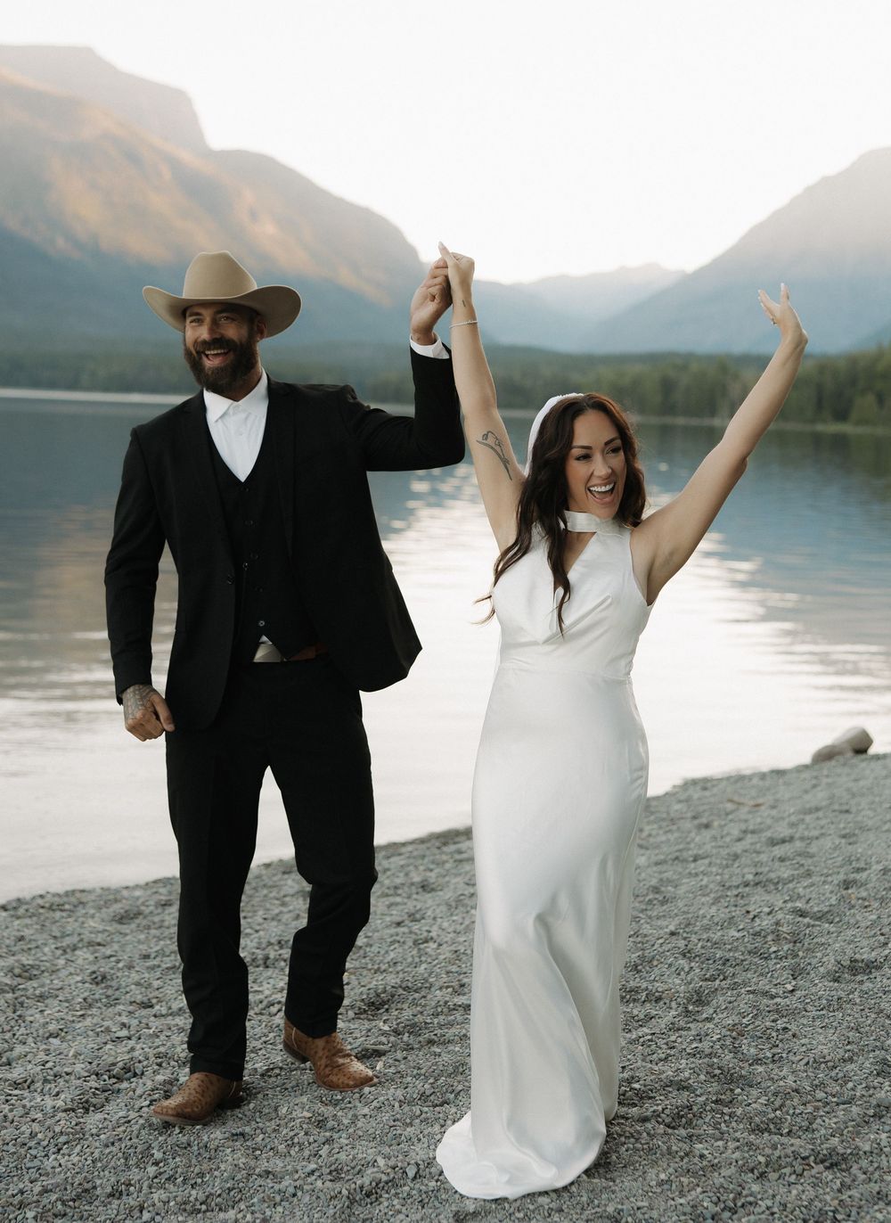 A joyful couple celebrates their wedding by a scenic mountain lake with the bride in white and groom in black.