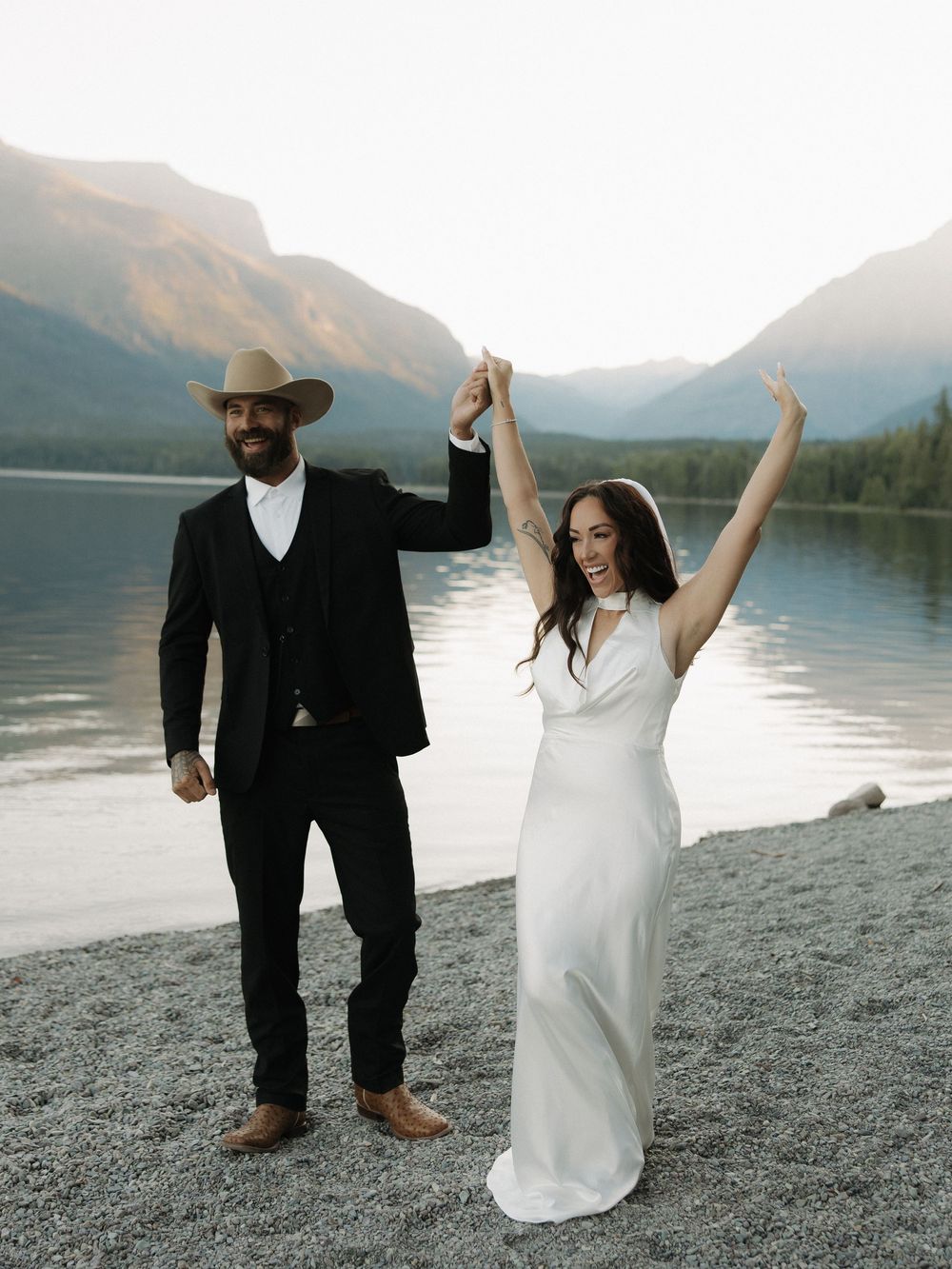 A joyful wedding couple dancing and celebrating on a rocky beach shore with mountains and lake in the background.