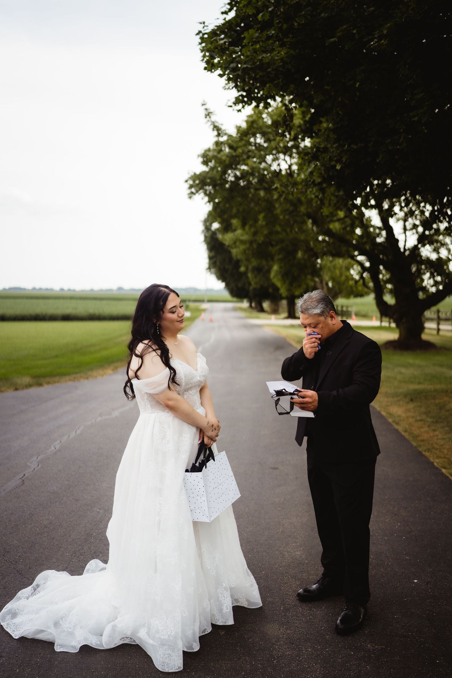 Two people sharing a light moment on a tree-lined country road, with one wearing a flowing white wedding gown.