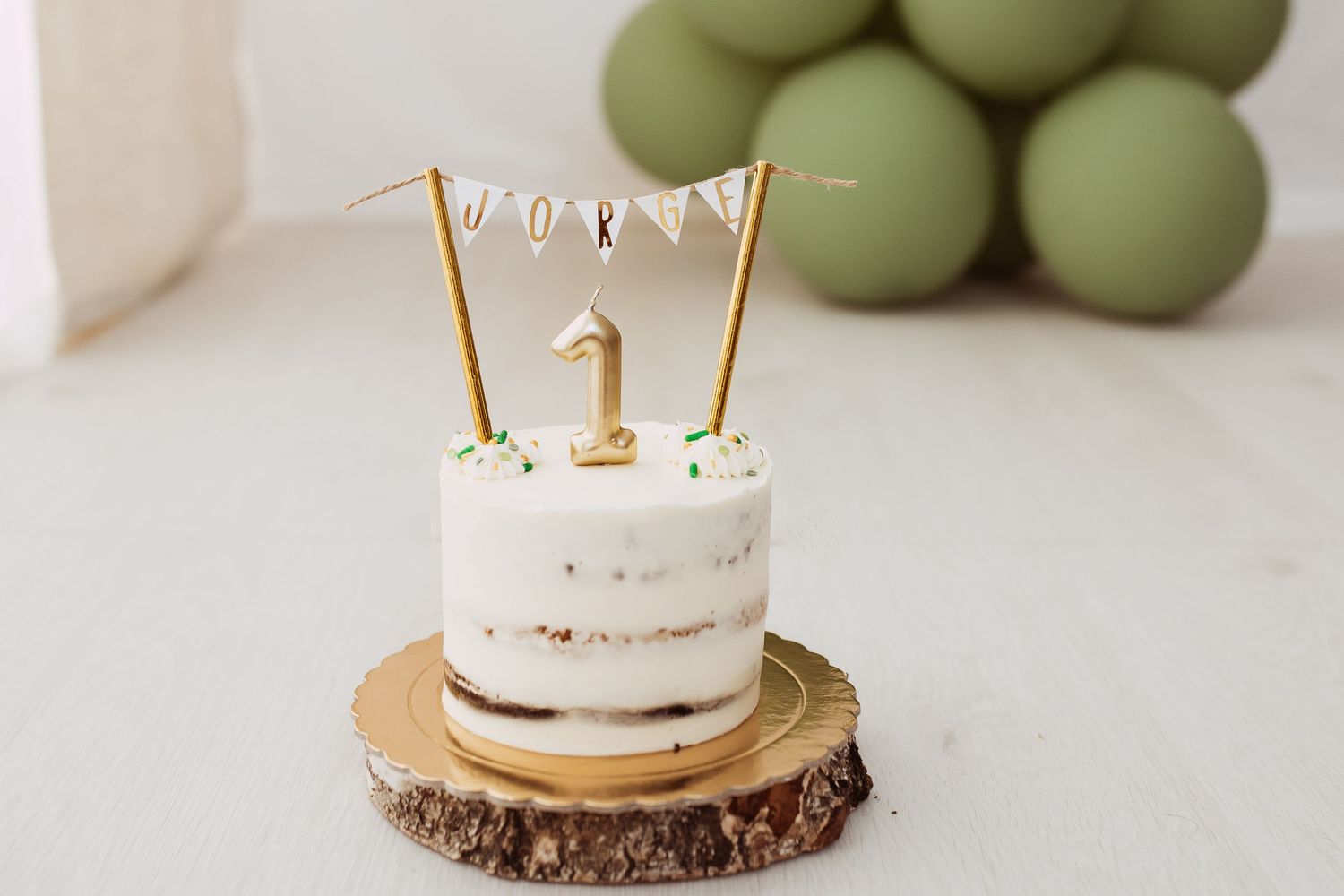Minimalist white cake with gold decorative toppers displayed on wooden base with green balloon backdrop.