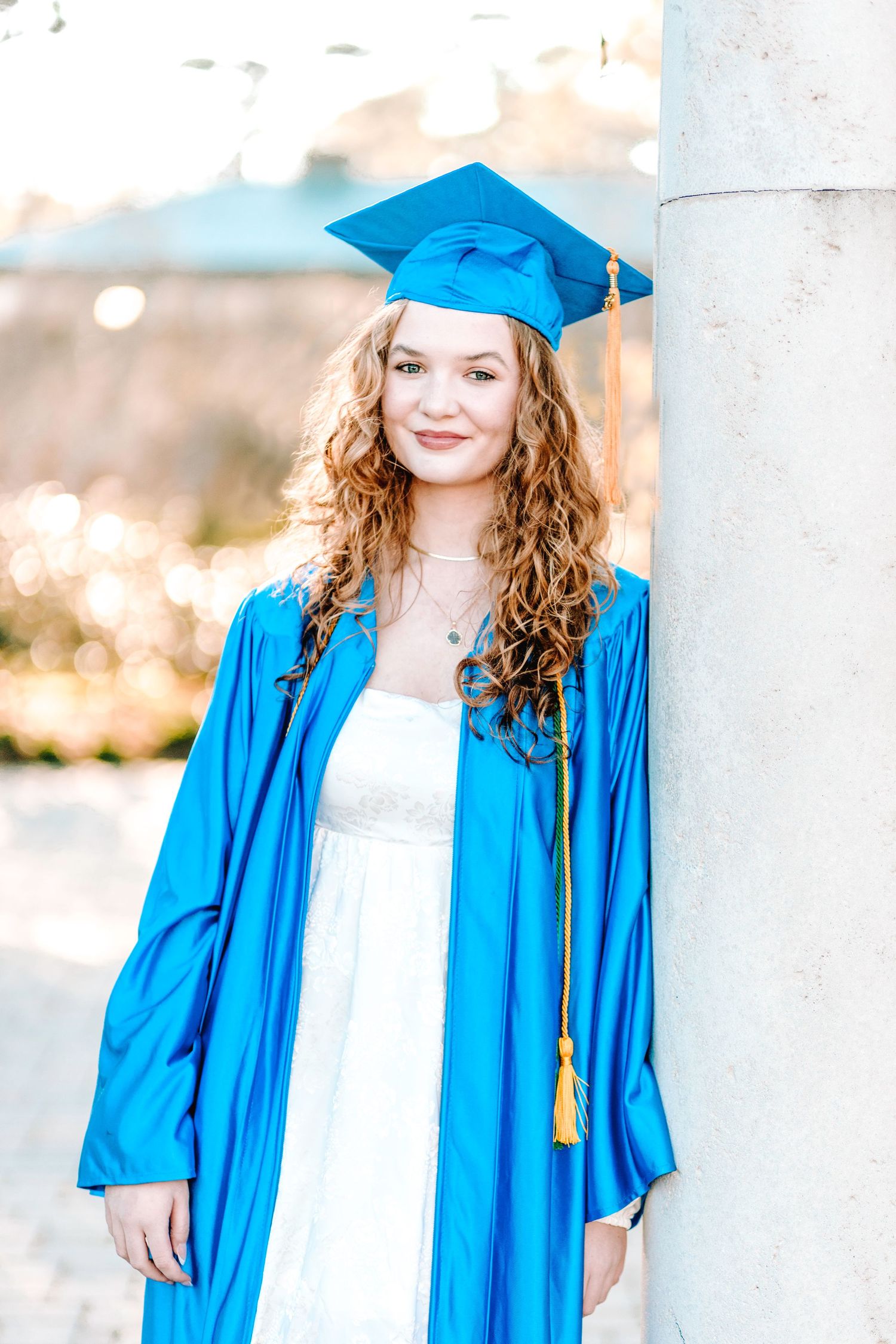 A graduate in a bright blue cap and gown stands outdoors against a blurred nature background.