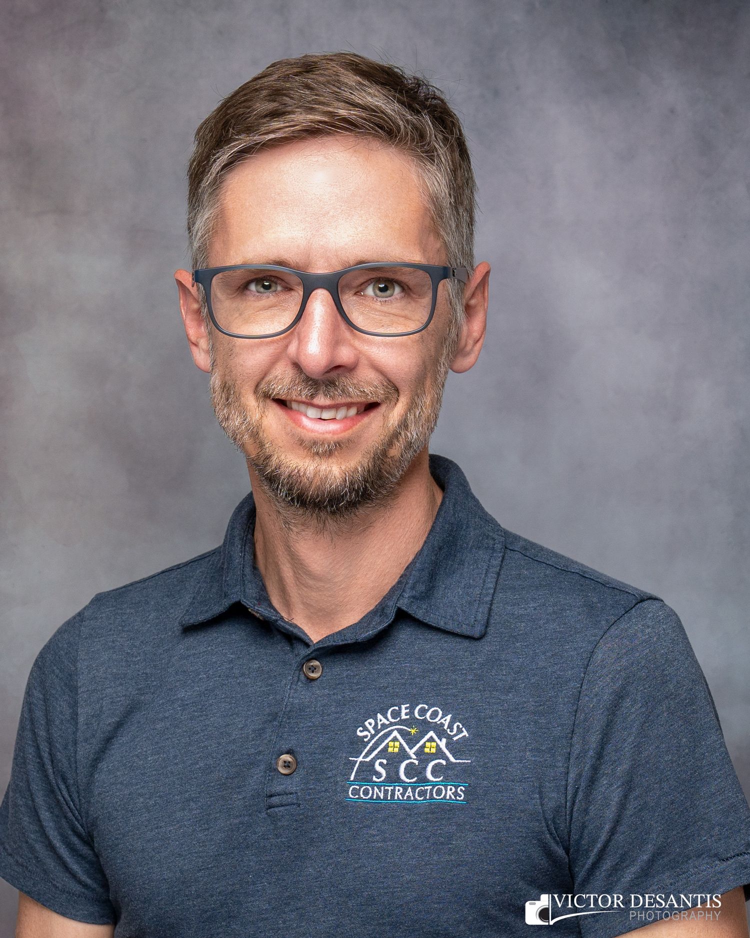 Professional headshot portrait showing a person in glasses and a navy polo shirt with company branding against a gray backdrop.