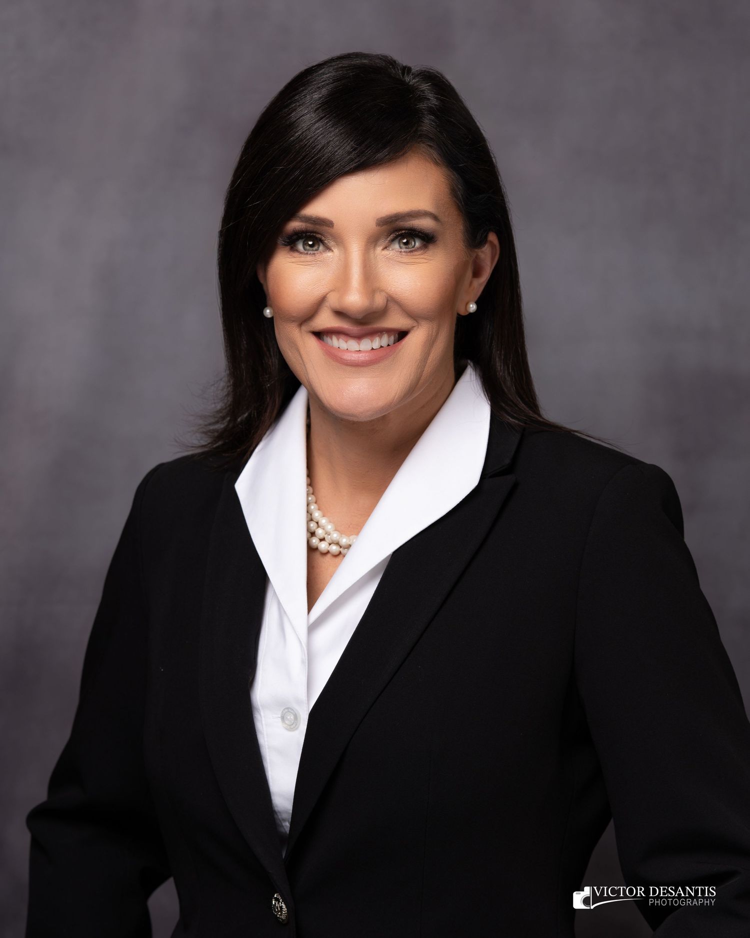 Professional business portrait of a smiling person in a black blazer and white blouse against a gray backdrop.