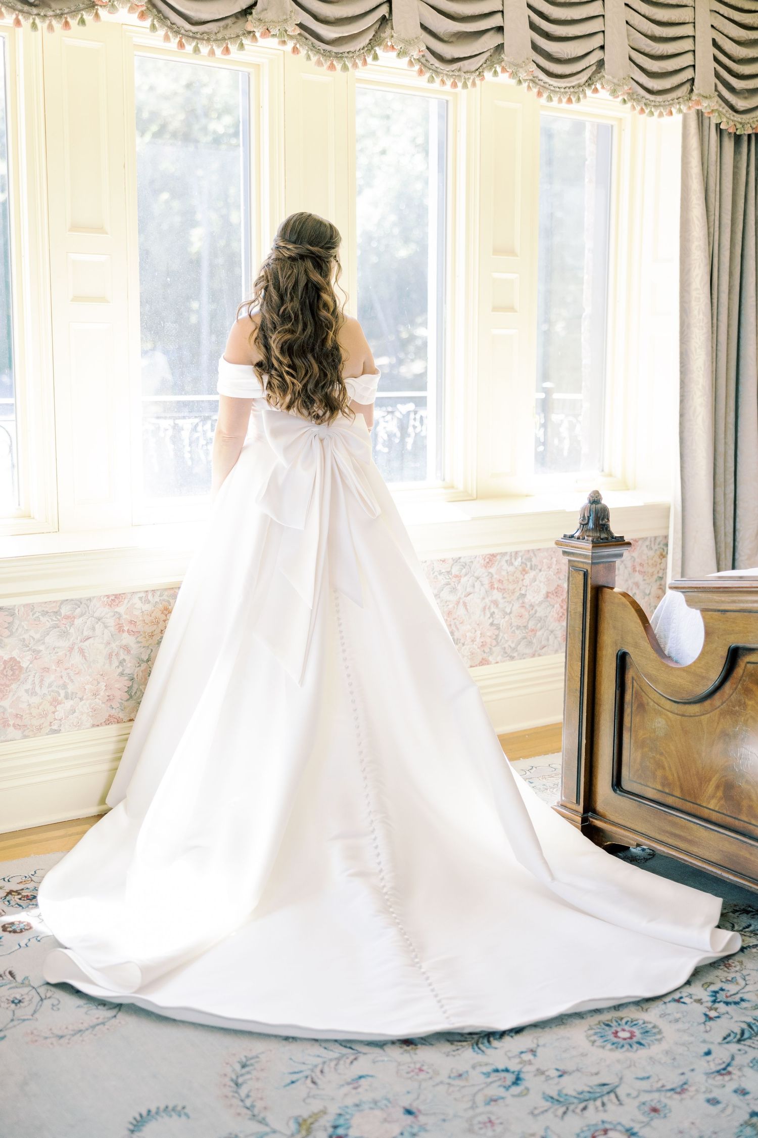 An elegant white wedding gown with a long train sweeps across a patterned carpet in a sunlit room.