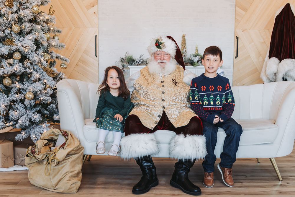 Children in holiday sweaters sit on a white couch near a decorated Christmas tree in a cozy living room setting.