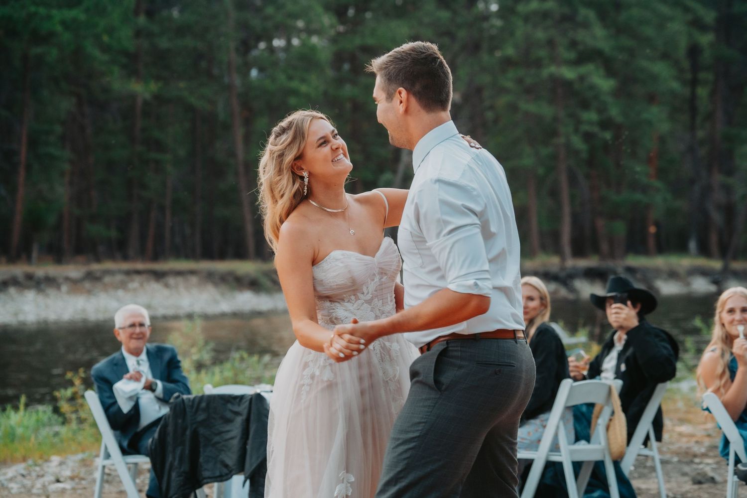 Couple shares their first dance at an outdoor forest wedding ceremony with guests seated on white chairs in the background.