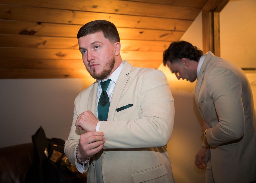Groomsman fixing tie and getting ready in warm-lit rustic room.