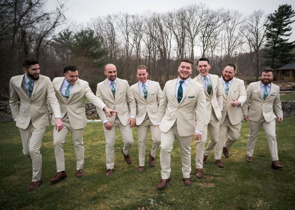 Groomsmen in cream suits pose playfully on a grassy field during wedding photos.