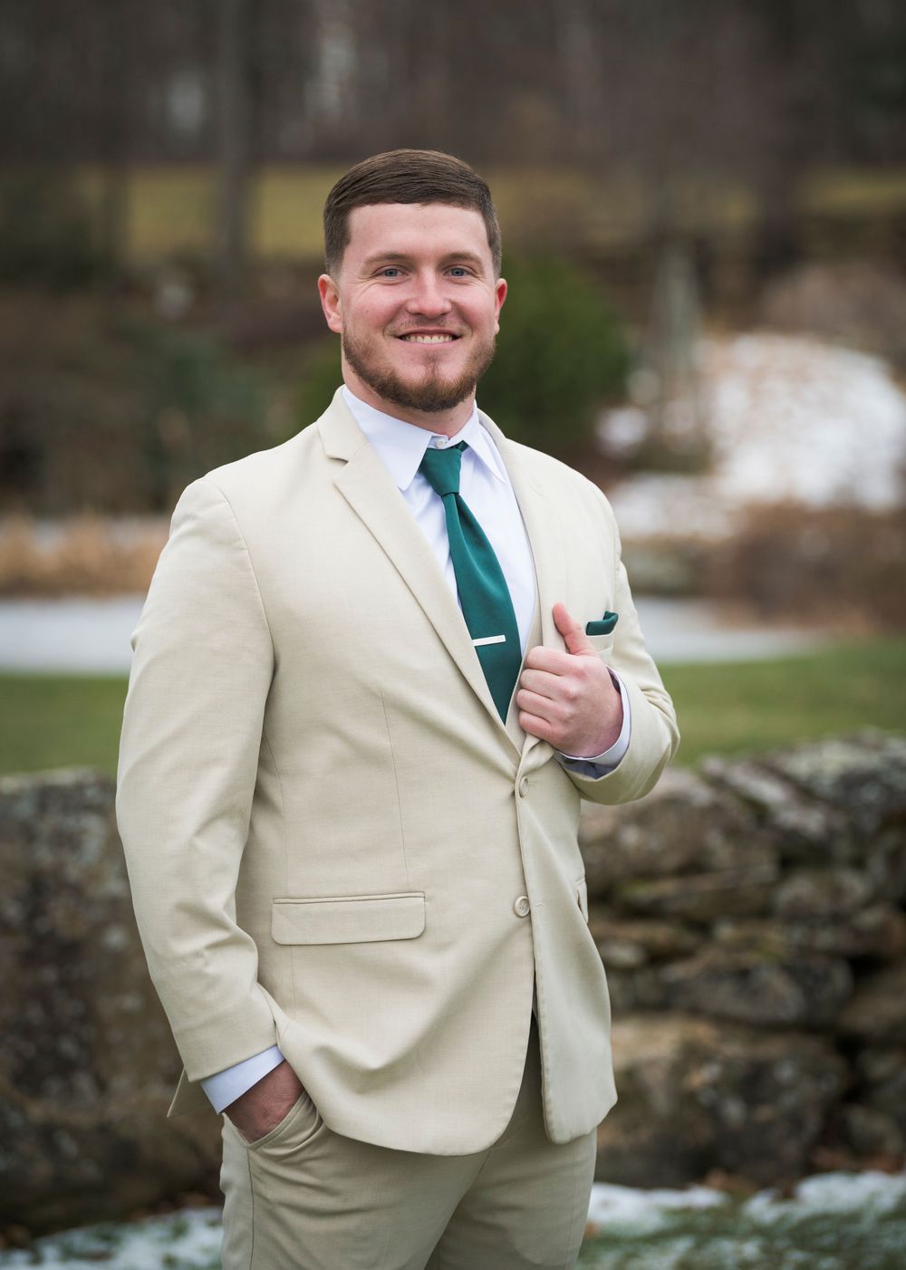 Groomsman in cream suit with teal tie poses confidently outdoors near stone wall.