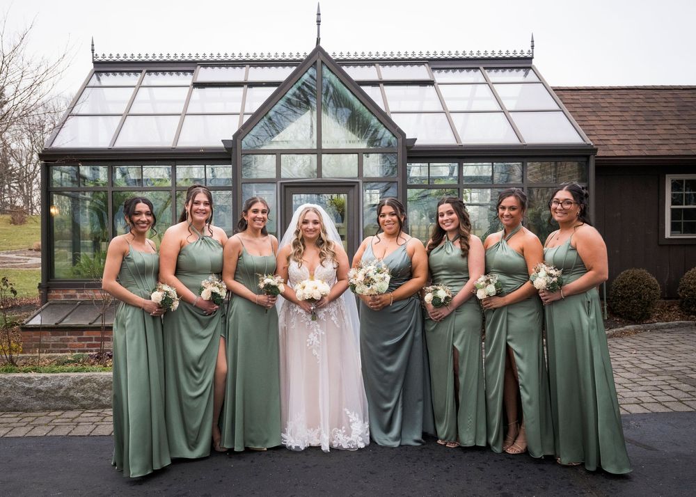 Wedding party in sage green dresses poses together outside a glass building.