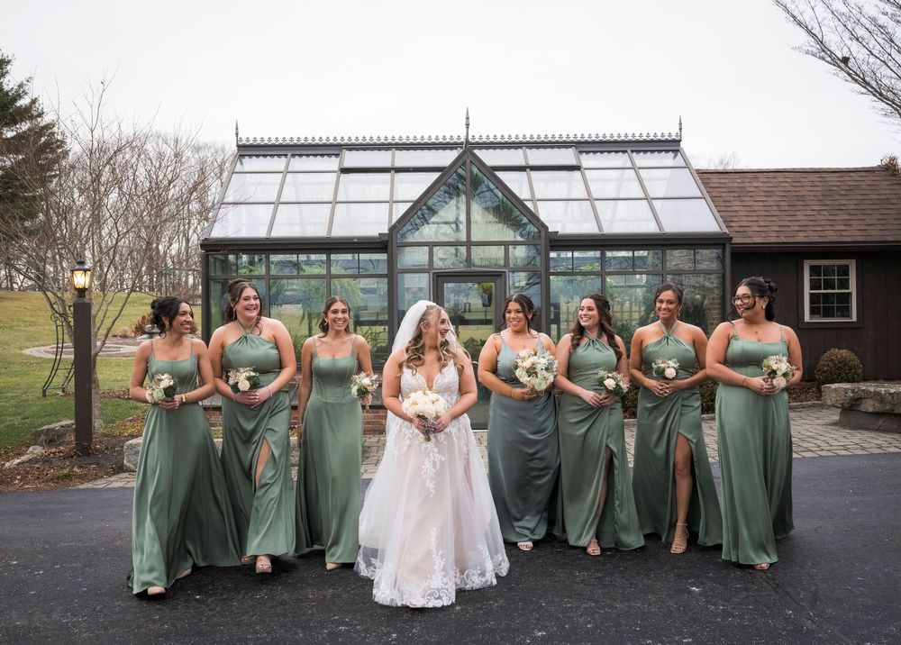 Wedding party in sage green bridesmaid dresses poses in front of a glass greenhouse conservatory.