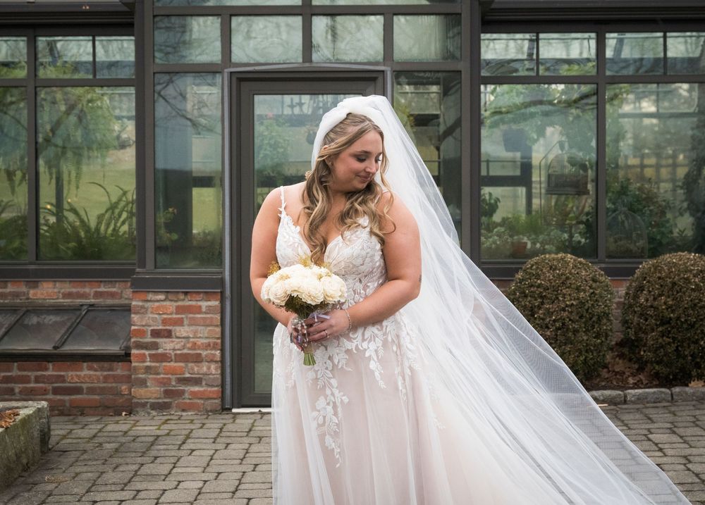 Bride in white wedding gown with flowing veil walking outside a brick building.