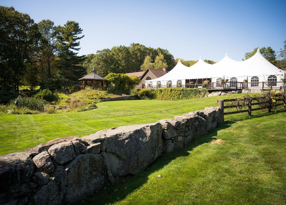 Stone wall lines a lush green lawn with white event tents and trees in the background on a sunny day.