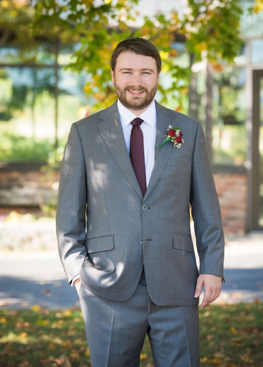 A groom in a gray suit with burgundy tie and boutonniere poses outdoors during autumn.