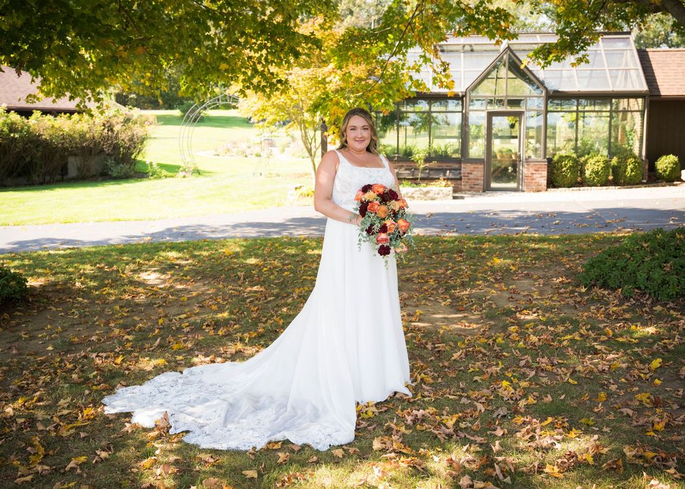A white wedding dress with lace details and flowing train photographed outdoors in a garden setting with a greenhouse.