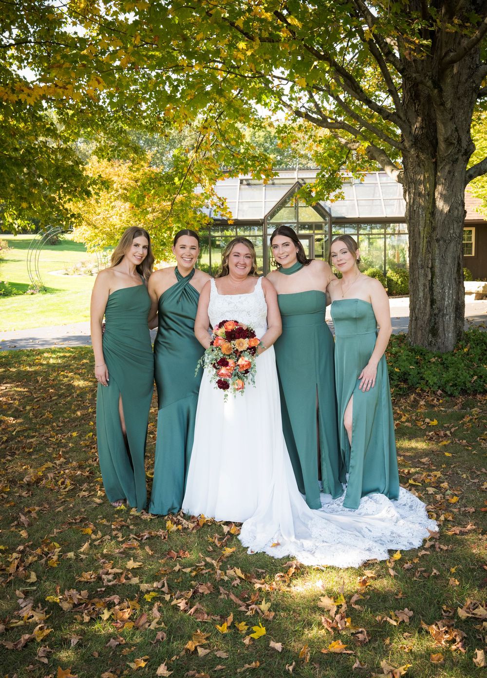 Wedding party poses outdoors in sage green dresses and white gown beneath autumn trees.