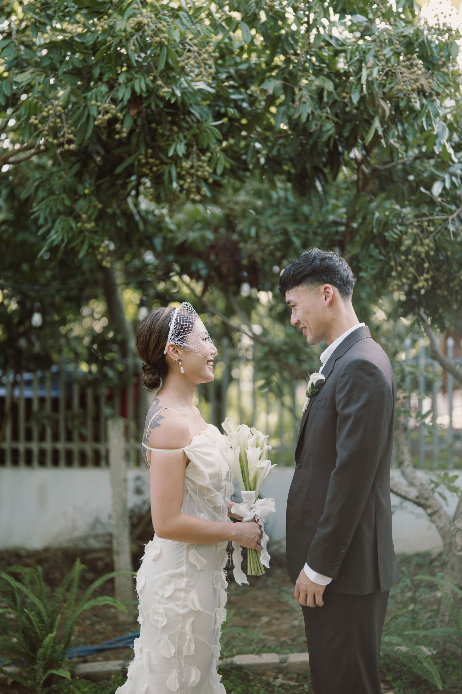 A couple in wedding attire shares a tender moment under green foliage during an outdoor ceremony.