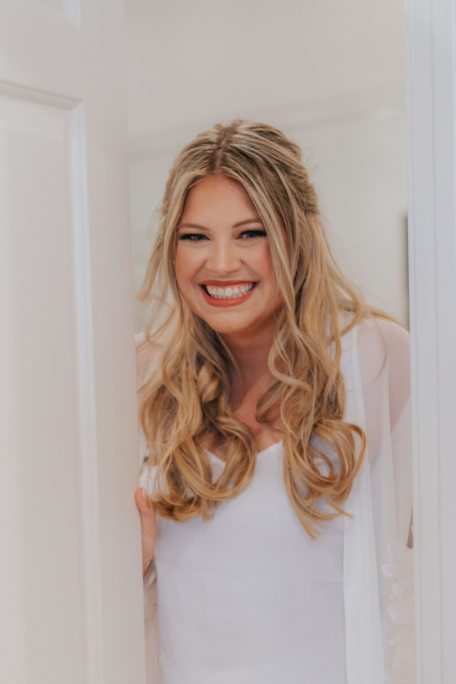 Woman with wavy blonde hair wearing a white top smiles brightly in a light-filled setting.