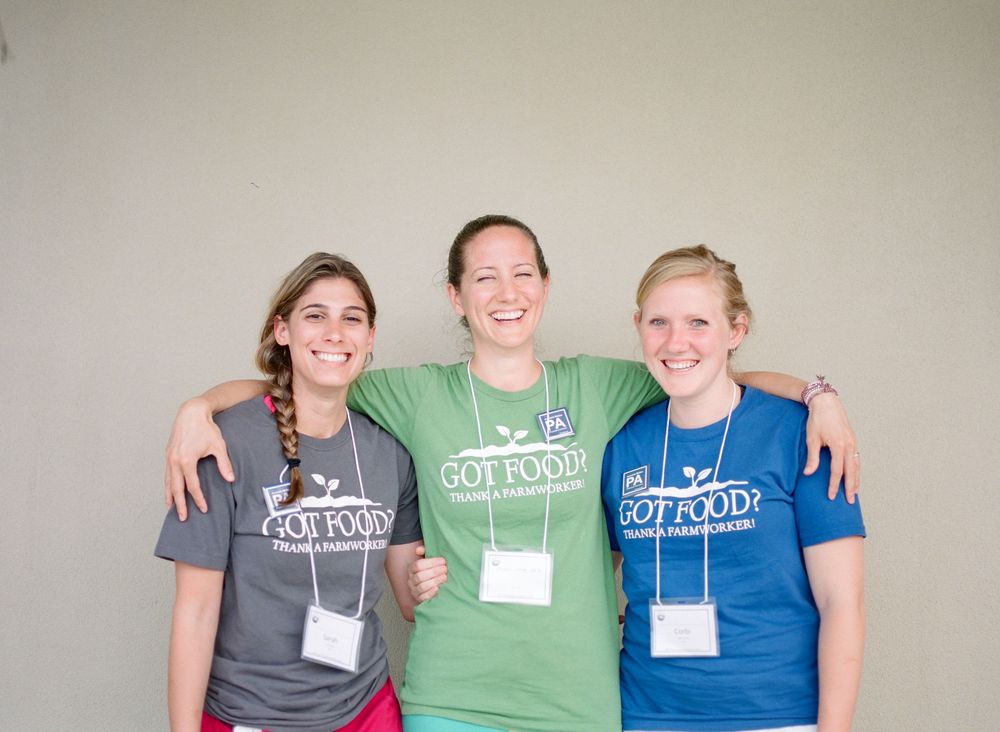 Three people wearing grey, green and blue branded t-shirts smile together against a neutral wall.