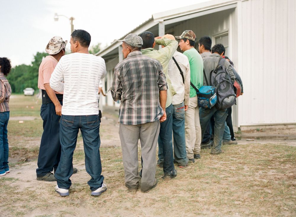 A line of people standing against a white building wall during an outdoor gathering.