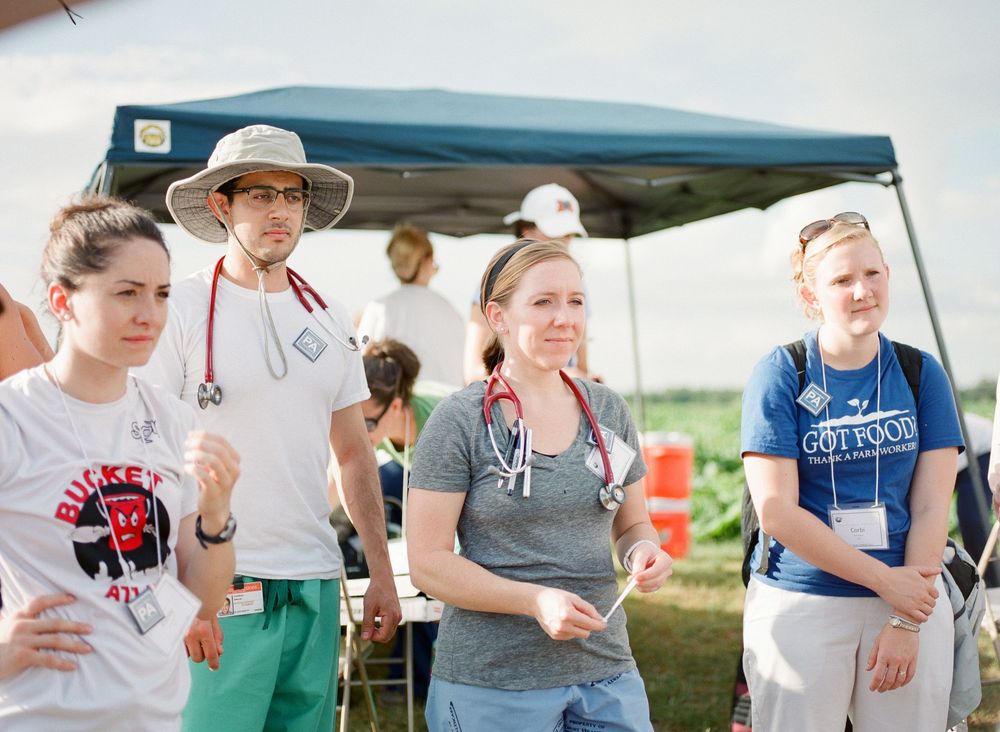 A group of people stand under a blue canopy tent at an outdoor event wearing casual summer clothing.