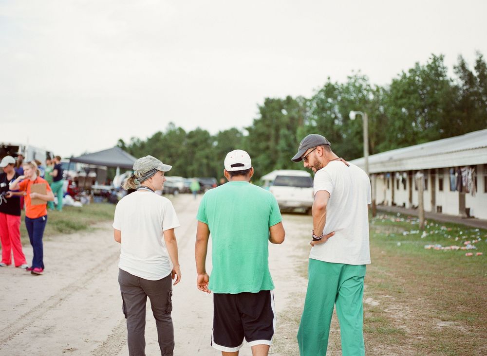Three people stand on a dirt path at an outdoor event with vendor tents and buildings visible in the background.