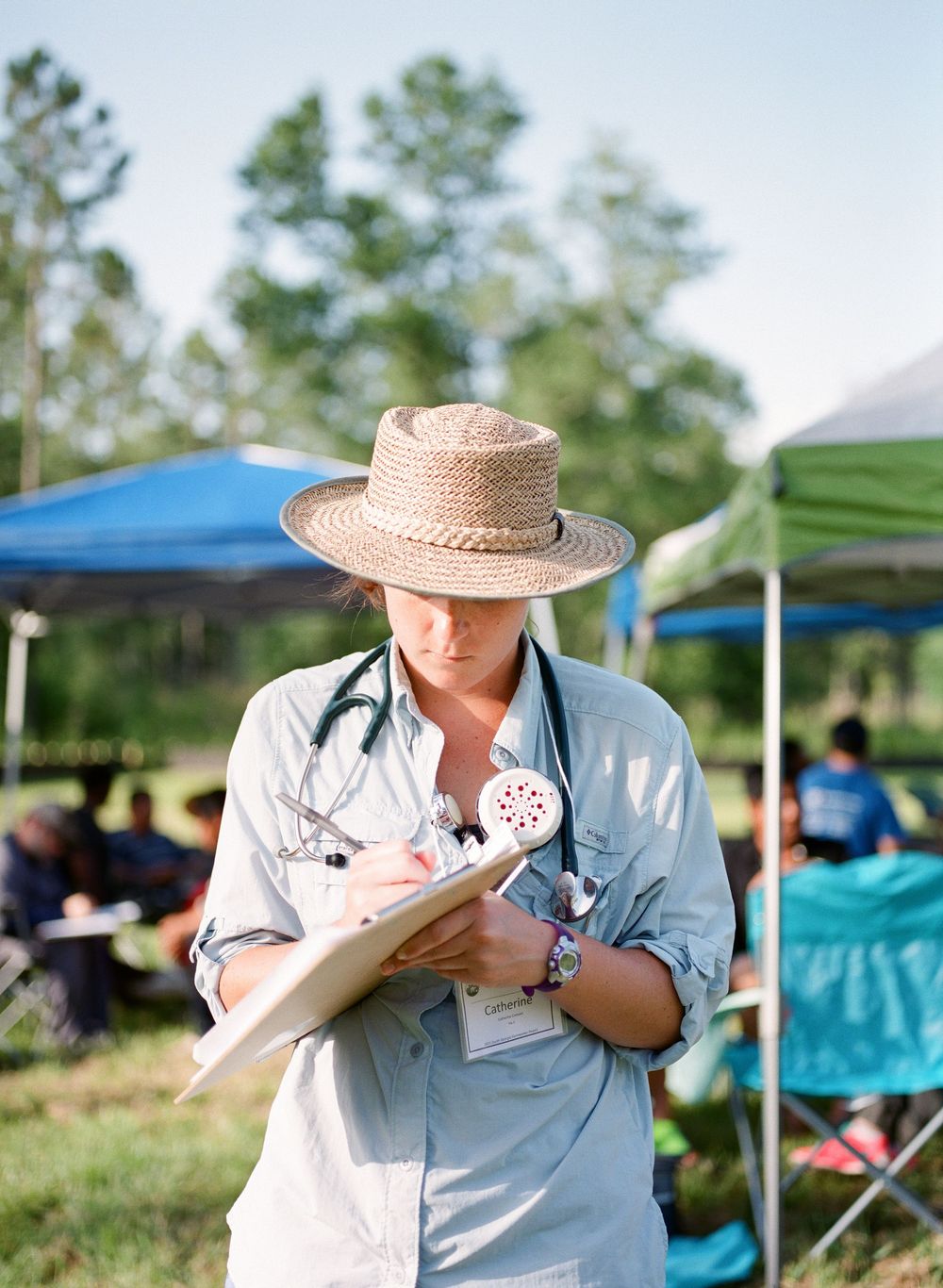 A person wearing a straw hat and light blue shirt takes notes during an outdoor event beneath canopy tents.