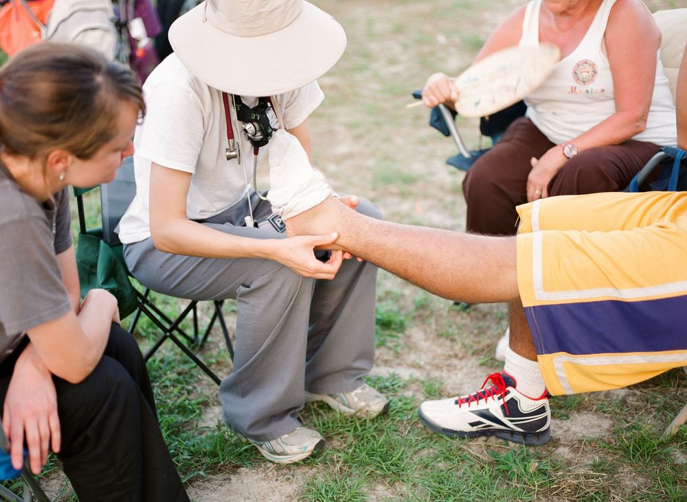 People sit in camping chairs on grass while treating and bandaging injuries at an outdoor first aid station.